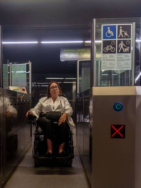 Kadın tekerlekli sandalye kullanıcısı bir metro istasyonunun ulaşılabilir kapısından çıkıyor, kentsel erişilebilirliği vurguluyor.