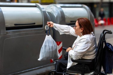 Elektrikli tekerlekli sandalyedeki kadın halka açık bir geri dönüşüm kutusu kullanıyor. Erişilebilirliği ve şehri kapsayıcılığı vurguluyor..