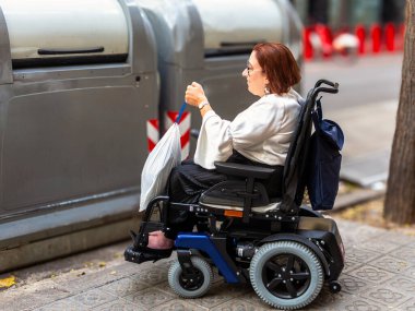 Elektrikli tekerlekli sandalyedeki bir kadın çöpü geri dönüşüm kutusuna atıyor, ulaşılabilir atık yönetimi çözümlerini gösteriyor.