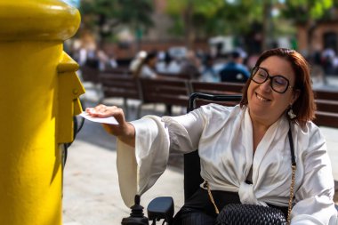 Kadın tekerlekli sandalye kullanıcısı sarı bir posta kutusuna şehir altyapısını ve erişilebilirliğini gösteren bir mektup gönderiyor..