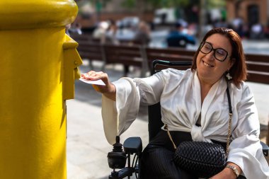 Tekerlekli sandalyedeki kadın, sarı bir posta kutusuna erişilebilirlik ve bağımsızlık gösteren bir mektup koyarken gülümsüyor..