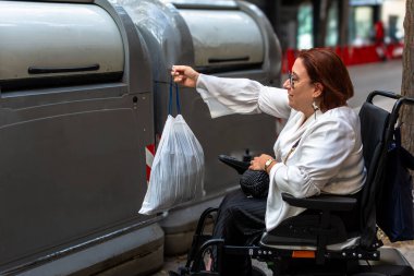 Tekerlekli sandalye kullanan bir kadın çöp torbasını halka açık bir geri dönüşüm noktasında bırakıyor, erişilebilirliği ve kapsayıcılığı teşvik ediyor..