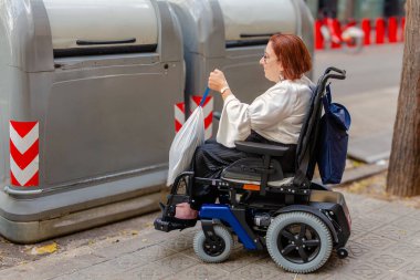 Kadın tekerlekli sandalye kullanıcısı çöpleri atmak, erişilebilirliği ve sürdürülebilirliği desteklemek için kentsel bir alanda geri dönüşüm kutusuna yaklaşıyor.