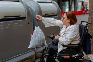 Kadın tekerlekli sandalye kullanıcısı çöp torbasını şehir geri dönüşüm kutusuna atıyor, kentsel erişilebilirliği vurguluyor.