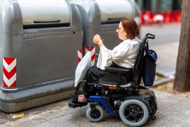 Tekerlekli sandalyedeki bir kadın, bir çöp torbasını geri dönüşüm kutusuna atar. Çevresel sorumluluğu ve ulaşılabilir şehir tesislerini teşvik eder..