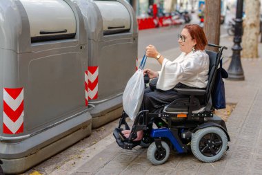 Kadın tekerlekli sandalye kullanıcısı bir çöp torbasını geri dönüşüm kutusuna koyuyor, bu da erişilebilir şehir hizmetleri ve çevre sorumluluğunu gösteriyor.