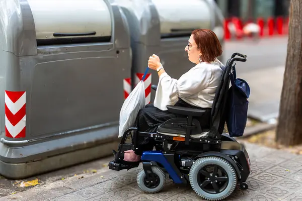 Tekerlekli sandalyedeki bir kadın, bir çöp torbasını geri dönüşüm kutusuna atar. Çevresel sorumluluğu ve ulaşılabilir şehir tesislerini teşvik eder..