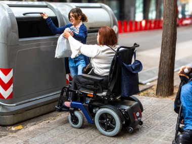 Tekerlekli sandalyedeki kadın kullanıcı, çöpleri geri dönüşüm kutusuna atarken, kentsel alanda erişilebilirliği ve desteği teşvik ederken yardım alıyor.