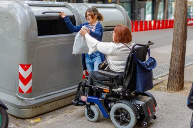 Tekerlekli sandalye kullanan bir kadın çöpü geri dönüşüm kutusuna atmak için başka bir kişiden yardım alıyor..