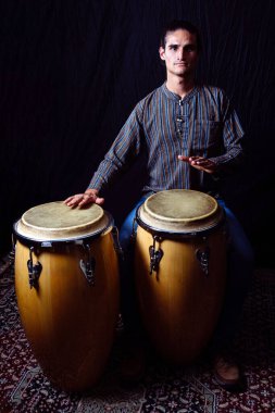 A male percussionist playing conga drums, captured in a full-body studio portrait. The dark backdrop contrasts with his traditional attire. clipart