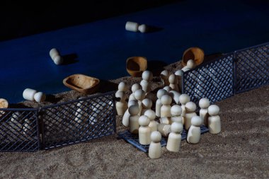 Wooden figurines and small boats scattered along a sandy shoreline confined by fences, symbolizing migration struggles and border challenges. clipart