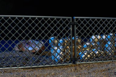 Clustered wooden figures behind a metal fence, illuminated by blue lighting, set on a textured sandy ground, representing themes of confinement. clipart