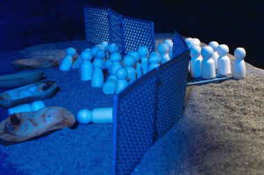 Abstract representation of migration struggles with wooden dolls in a sandy area, separated by a fence under blue lighting. clipart