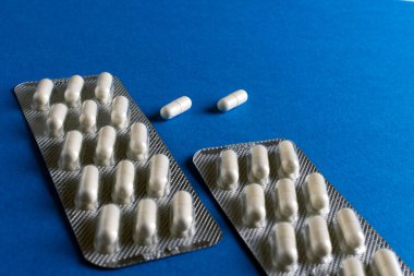 İki beyaz kapsül paketi, biri açık, gevşek haplar etrafa saçılmış, parlak mavi bir arka planda sergileniyor..