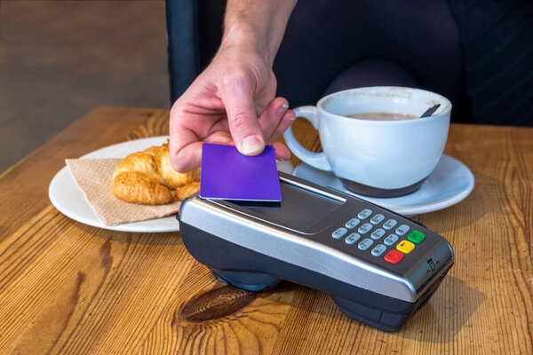 A closeup of a womans hand holding a purple card while paying with a contactless terminal at a cafe table featuring a croissant and coffee.
