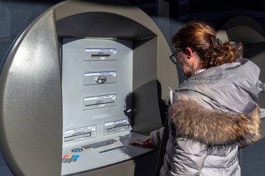 Gözlüklü ve kışlık ceketli bir kadın açık hava güneşli bir günde ATM makinesine ödeme kartı yerleştiriyor.