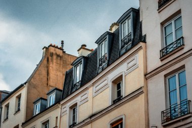 Yumuşak pastel duvarları, karmaşık pencereleri ve bulutlu gökyüzüne karşı geleneksel mansard çatıları olan klasik Paris binaları..
