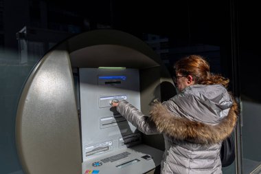 Kışlık ceketli bir kadın güneşli bir günde şehir merkezinde açık hava ATM 'sine kart sokuyor..