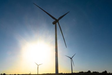 Bir rüzgar türbini, arkasında güneş parıldarken dik durur ve dramatik bir siluet yaratır. Akşam karanlığında yenilenebilir enerji manzarasını tamamlayan iki türbin daha..