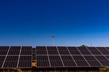 Fotovoltaik güneş panelleri, açık mavi gökyüzünün altında bir iletişim kulesi ile yenilenebilir enerji üretiyor..