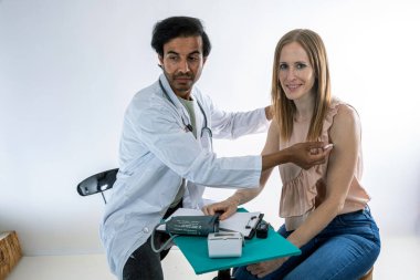 Doktor, aşıyı yaptıktan sonra kadın bir hastanın koluna yapışkan bandaj uyguluyor, tıbbi bir ortamda aşıyı gösteriyor, masada tıbbi ekipmanlar var.