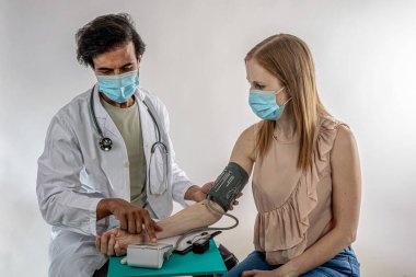 Bir kadın hastanın kan basıncını ölçen yüz maskesi takan bir doktor aynı zamanda klinikteki tıbbi muayene sırasında maske takarak sağlık hizmetlerinde güvenlik ve bakımı sağlıyordu.