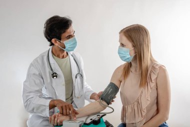 Laboratuvar önlüğü ve steteskop giyen doktor, hastanede veya klinikte yapılan muayene sırasında her ikisi de koruyucu maske takan kadın hastanın kan basıncını ölçüyor.