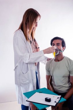 Laboratuvar önlüğü ve steteskop takan kadın doktor tıbbi bir ortamda erkek bir hastaya oksijen maskesi takıyor. Yanında bir pano, nabız oksimetresi ve kalem var.