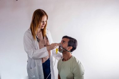 Laboratuvar önlüğü ve steteskop giyen kadın doktor hastane ortamında bir erkek hastaya oksijen maskesi takıyor ve solunum desteği ve bakım sağlıyor.