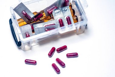 Kırmızı haplar su toplayan bir paketten ve şeffaf bir plastik kutudan beyaz bir yüzeye dökülüyor. Bu da ilaç, sağlık ve ilaç ürünleri temaları olduğunu gösteriyor.
