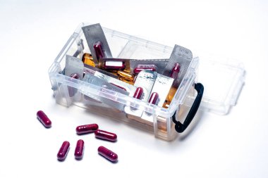 Şeffaf plastik organizatör kutusu çeşitli haplar, kapsüller ve su toplayıcılarıyla dolup taşıyor, bazıları beyaz yüzeye dökülüyor, ilaçların bolluğunu ve düzensizliğini vurguluyor