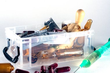 Ahşap manken, ilaç, kapsül ve ampullerle dolu saydam bir kutuda, uyuşturucu bağımlılığı ve aşırı doz temalarını gösteren bir şırınganın yanında yatıyor.