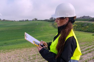 Güvenlik kaskı ve yelek giyen kadın mühendis, bir panoya not alıyor ve rüzgar türbini çiftliğinde akıllı telefon kullanıyor, yenilenebilir enerji geliştirme üzerinde çalışıyor.