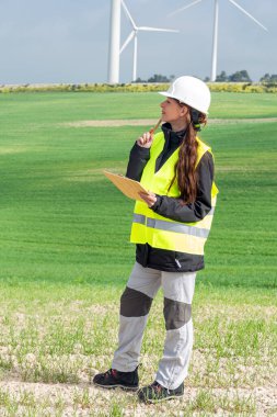 Güvenlik kaskı ve yelek giyen kadın mühendis elinde pano ve kalem, yeşil bir alanda rüzgar türbinlerini denetliyor, sürdürülebilir enerji üretimi planlıyor.