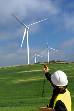 Bir anemometre ve pano tutan kadın mühendis rüzgar hızını ölçüyor ve sürdürülebilir bir enerji çiftliğinde rüzgar türbinlerinin yanında yenilenebilir enerji üretimi için veri topluyor.
