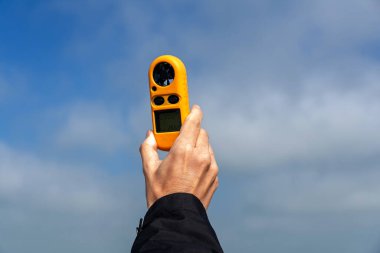 Rüzgarın bulutlu gökyüzüne karşı hızını ölçen bir anemometre tutan, rüzgar enerjisi üretiminin potansiyelini değerlendiren ve sürdürülebilir uygulamaları destekleyen bir el.