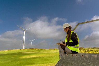 Güvenlik yeleği ve kask giyen kadın mühendis yeşil bir alanda rüzgar türbinlerini incelerken not alıyor. Mavi gökyüzü altında sürdürülebilir enerji gelişimine katkıda bulunuyor.