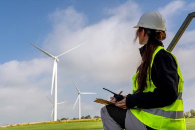 Güvenlik yeleği ve miğfer takan kadın mühendis ellerinde telsiz ve bir alandaki rüzgar türbinlerini inceleyerek yenilenebilir enerji ve sürdürülebilir gelişime katkıda bulunuyor.