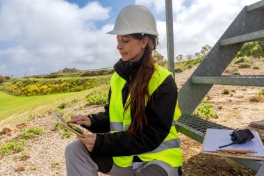 Güvenlik kaskı ve yeleği takan kadın mühendis, yenilenebilir enerji sağlamak için dijital tablet kullanırken rüzgar türbini bölgesinde metal bir merdivenin yanında çömeliyor.