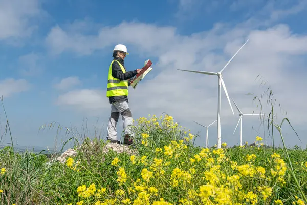 Çelik yelek ve kask giyen kadın mühendis çiçek açan bir alanda rüzgar türbinlerinin yakınındaki planları inceliyor, sürdürülebilir enerji gelişimi ve çevresel sorumluluğu temsil ediyor.