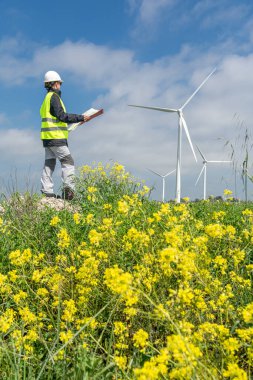 Güvenlik kaskı ve yelek giyen kadın mühendis, sarı çiçeklerden oluşan bir alanda rüzgar türbinlerini incelerken, sürdürülebilir ve yenilenebilir enerji üretimini destekliyor.