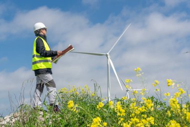 Güvenlik kaskı ve yelek giyen kadın mühendis, çiçek açan bir alanda rüzgar türbinlerinin yakınında, sürdürülebilir enerji ve ekolojik farkındalığı destekliyor.