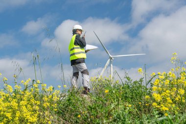 Güvenlik yeleği ve miğfer takan kadın mühendis, arka planda sarı çiçekler ve mavi gökyüzü olan rüzgar türbinindeki belgeleri incelerken telsiz kullanıyor.