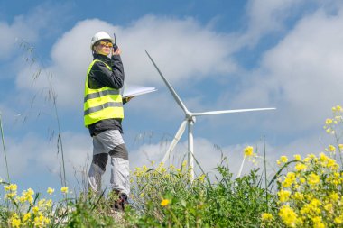 Güvenlik kaskı ve yeleği takan kadın mühendis, telsiz kullanıyor ve sarı çiçek tarlasında rüzgar türbinlerini incelerken elinde belgeler tutuyor, sürdürülebilir enerji oluşturuyor.