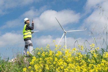 Güvenlik kaskı ve yelek giyen kadın mühendis telsiz ve tablet kullanarak mavi gökyüzünün altındaki bir rüzgar türbininin yanındaki sarı çiçek tarlasında duruyor.