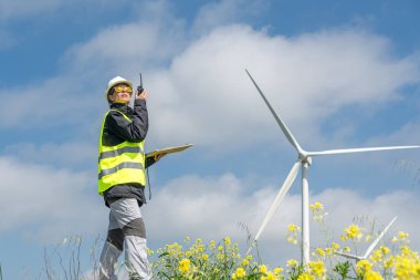 Güvenlik yeleği ve kask takan kadın mühendis, bulutlu mavi gökyüzünün altındaki bir rüzgar türbini çiftliğinde telsiz ve pano ile operasyonları yönetiyor.