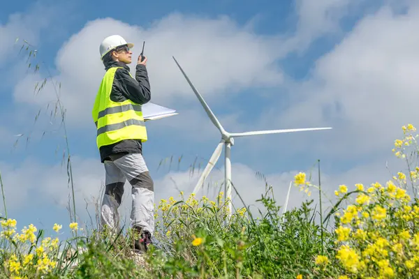 Güvenlik yeleği ve miğfer takan kadın mühendis, mavi gökyüzü altında sürdürülebilir ve yenilenebilir enerji üreten rüzgar türbini santralinin planlarını tutarken telsiz kullanıyor.
