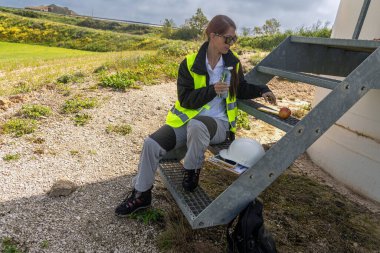 Yüksek görüş mesafesi olan bir ceketli kadın mühendis rüzgar tarlasında metal merdivenlerde elinde aperatifle oturuyor. Sürdürülebilir enerji sektörlerinde kadınların rolünü temsil ediyor.