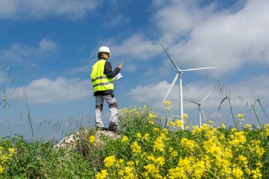 Güvenlik yeleği ve kask giyen kadın mühendis anemometre kullanıyor ve sarı çiçeklerden oluşan bir alanda rüzgar türbinlerini incelerken notlar alıyor, sürdürülebilir enerji gelişimini destekliyor.