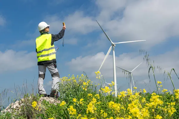 Yelek ve miğfer takan mühendis rüzgâr hızını sarı çiçeklerden oluşan bir alanda ölçerek sürdürülebilir enerji için rüzgar türbinlerinin verimliliğini hesaplıyor.